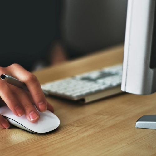 A hand using a wireless mouse at a modern desk setup with a computer and keyboard.