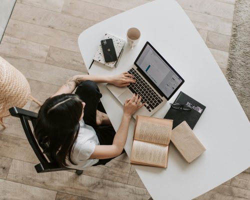 Overhead view of a woman using a laptop at a home desk, surrounded by books, a phone, and a cup.